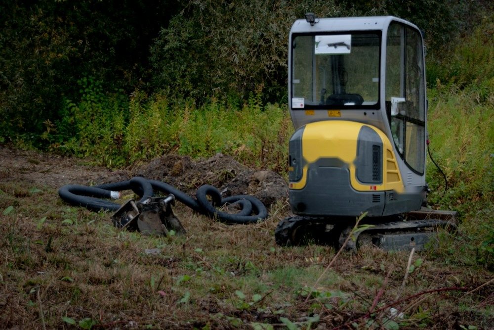 A mini excavator pulling stumps and brush on a partly cleared wooded lot
