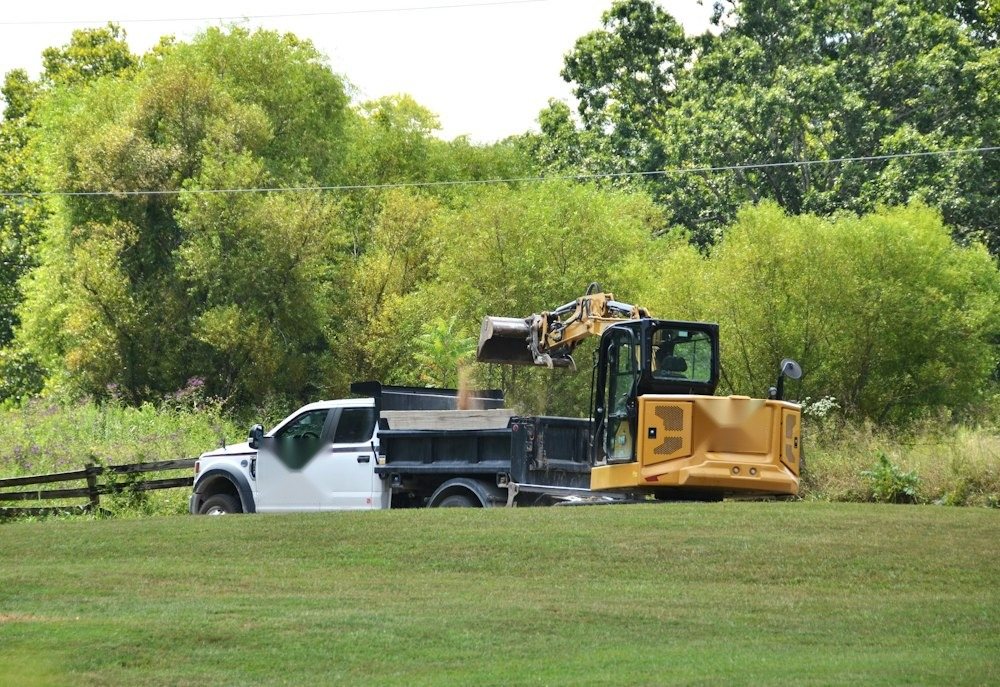 A compact track loader parked at the edge of a field beside a tree line